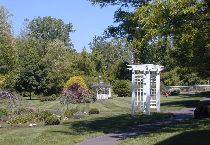 Manicured garden with a white arbor, a gazebo, walkways and trees under a clear blue sky.