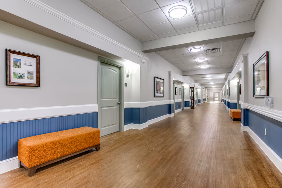 A long, well-lit hallway in a senior living facility with wood flooring, light gray walls with blue wainscoting, framed pictures on the walls, and orange cushioned benches placed along the corridor.