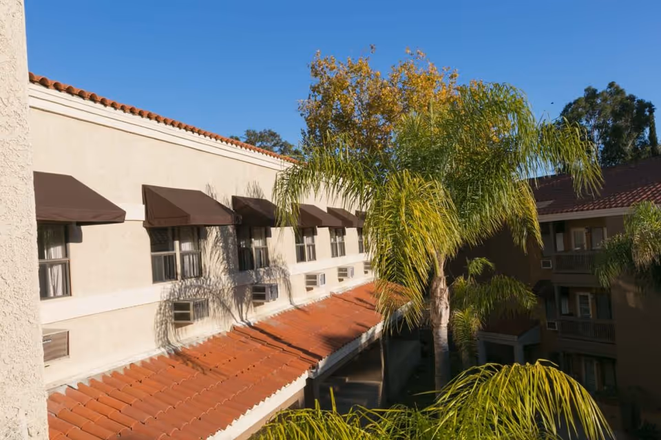 Exterior view of a multi-story building with beige walls, brown window awnings, and red tile roofing. Palm trees and other greenery are visible in the foreground under a clear blue sky.