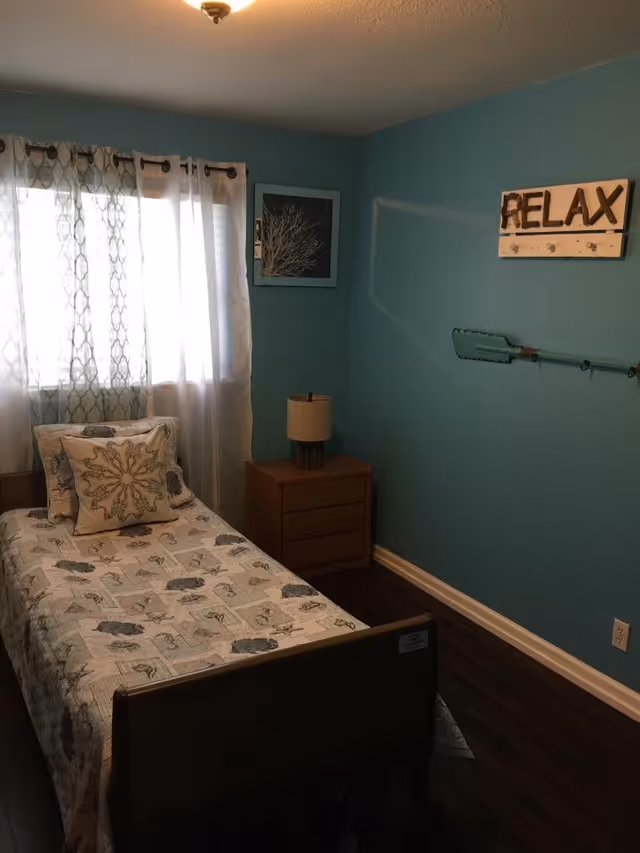 A small bedroom with teal walls, a single bed with a patterned bedspread and pillows, a wooden nightstand with a lamp, a window with sheer and patterned curtains, a framed picture on the wall, and a wall decoration with the word 'RELAX'.