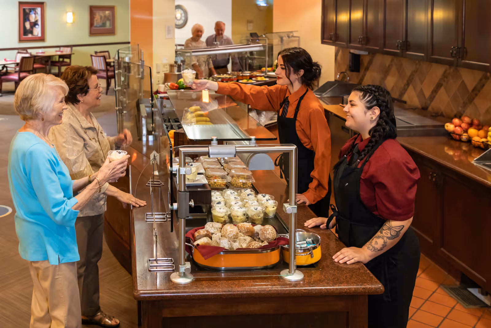 Two servers behind a buffet counter handing desserts to smiling elderly residents in a senior living dining area.