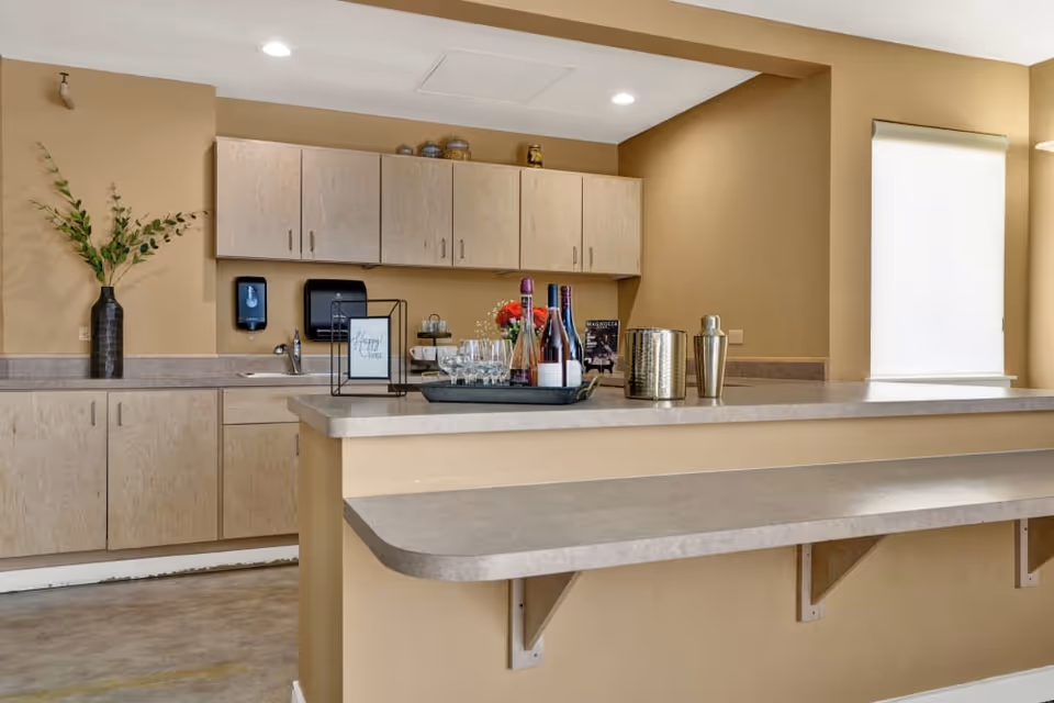 Interior view of a kitchenette area with beige walls and light wood cabinets. A countertop bar with a tray holding wine bottles, glasses, and a shaker is in the foreground. A vase with green foliage is on the left side of the counter, and a window with a white roller shade is on the right.