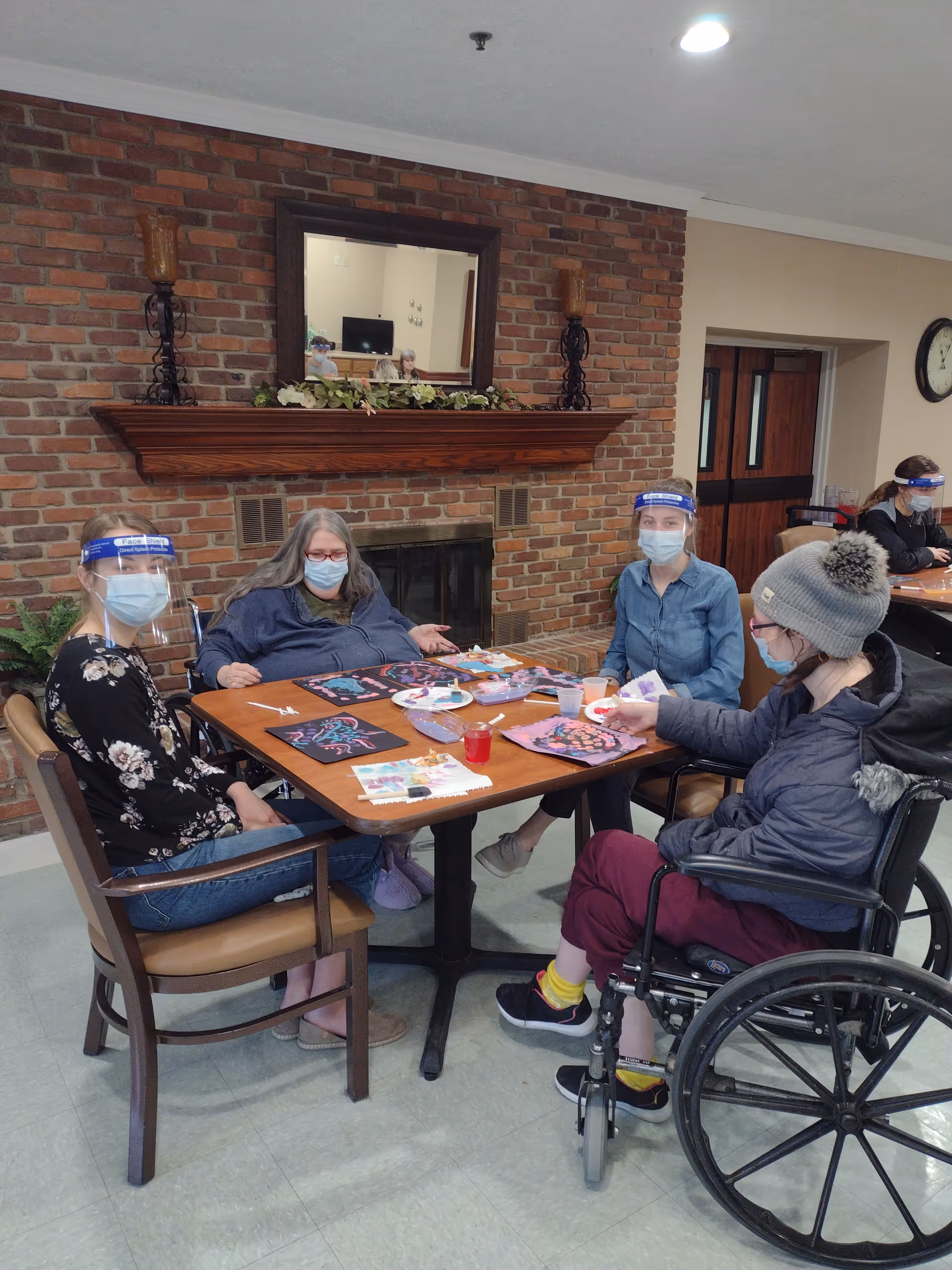 Four people wearing face masks and face shields sitting around a table in a room with a brick fireplace. They are engaged in an art activity with paint and paper. One person is in a wheelchair. The room has a mirror above the fireplace and a clock on the wall.