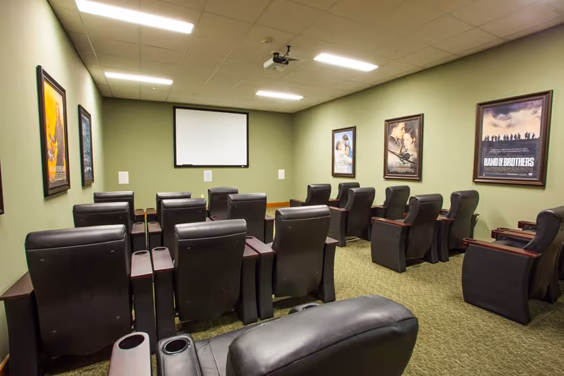 A small movie theater room with multiple rows of black leather recliner chairs facing a blank projector screen on a green wall. Movie posters are framed and hung on the walls, and the ceiling has recessed lighting and a mounted projector.