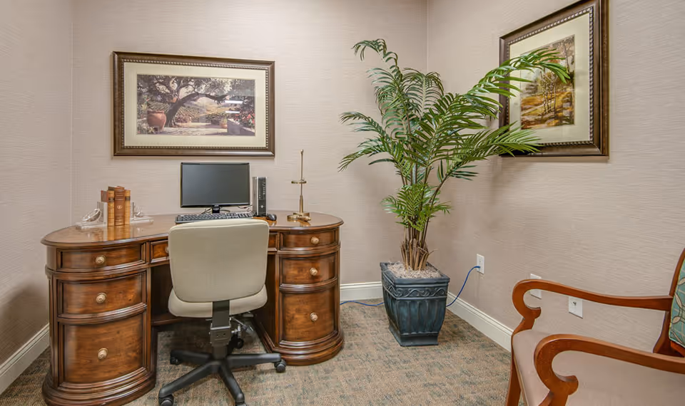A small office corner in a senior living facility featuring a wooden desk with multiple drawers, a computer monitor, keyboard, and mouse. A beige office chair is positioned at the desk. There are two framed landscape paintings on the beige walls and a large potted plant in a decorative blue planter. A wooden armchair with a patterned cushion is partially visible on the right side.