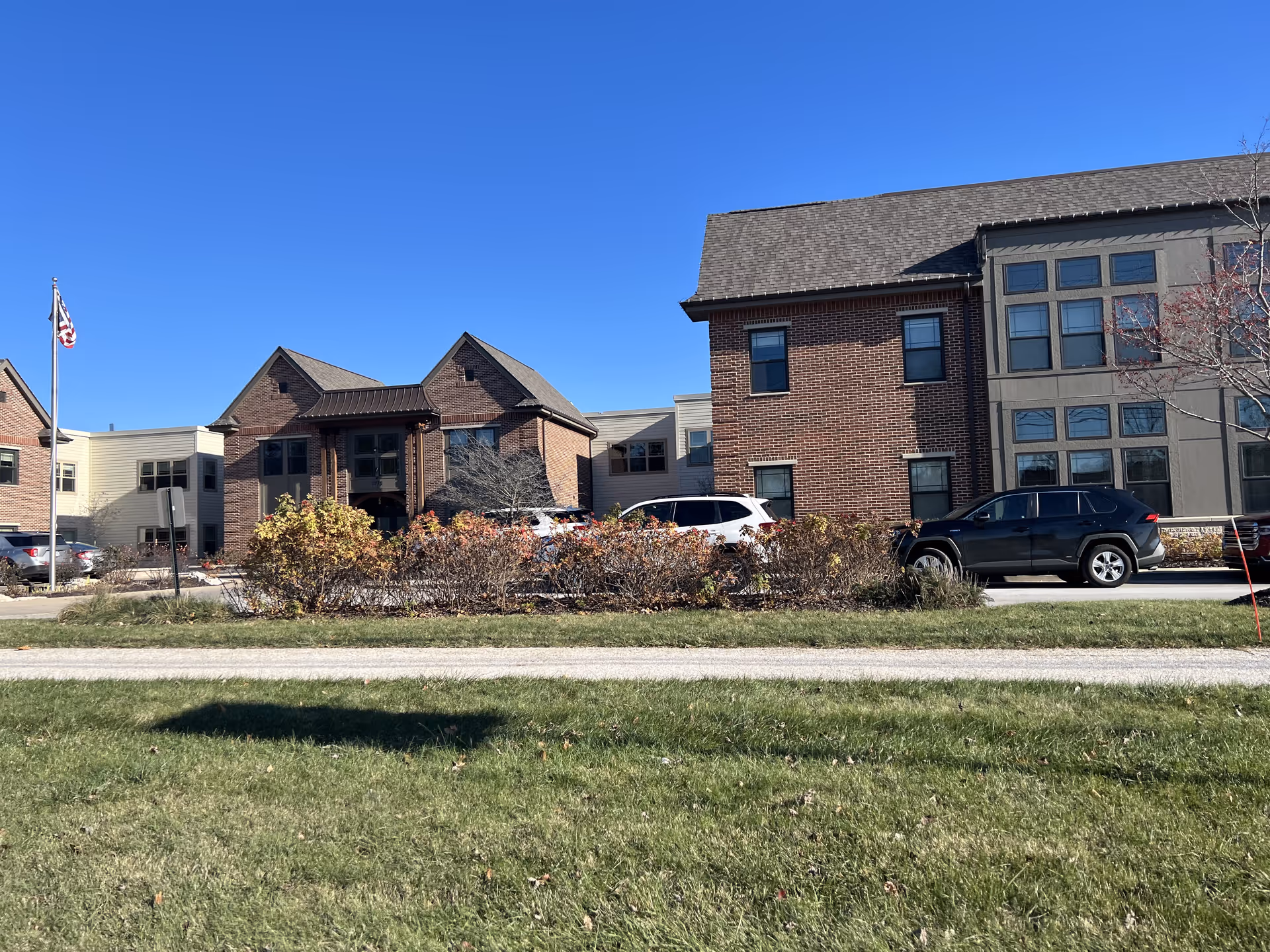 Exterior view of a senior living facility building with brick and beige siding, several windows, parked cars, a flagpole with an American flag, and landscaping with bushes and grass under a clear blue sky.