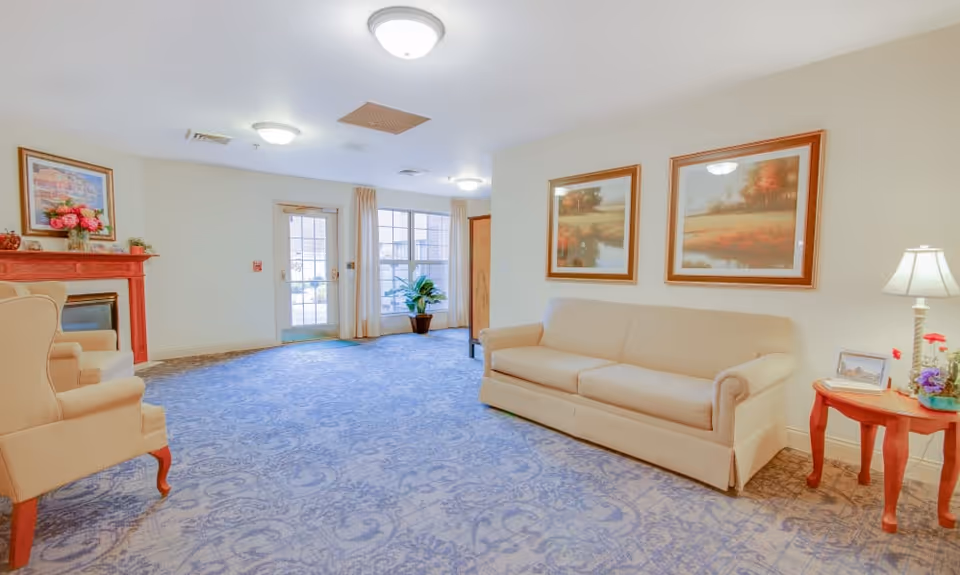 A bright and spacious living room area with a beige sofa and two beige armchairs. The room has a blue patterned carpet, a red wooden fireplace with a floral arrangement on top, two framed landscape paintings on the wall above the sofa, a small wooden side table with a lamp and flowers, and large windows with cream curtains letting in natural light.