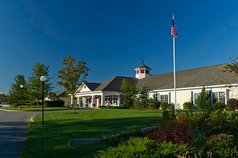 Single-story senior living building with a flagpole and landscaped lawn under a clear blue sky.