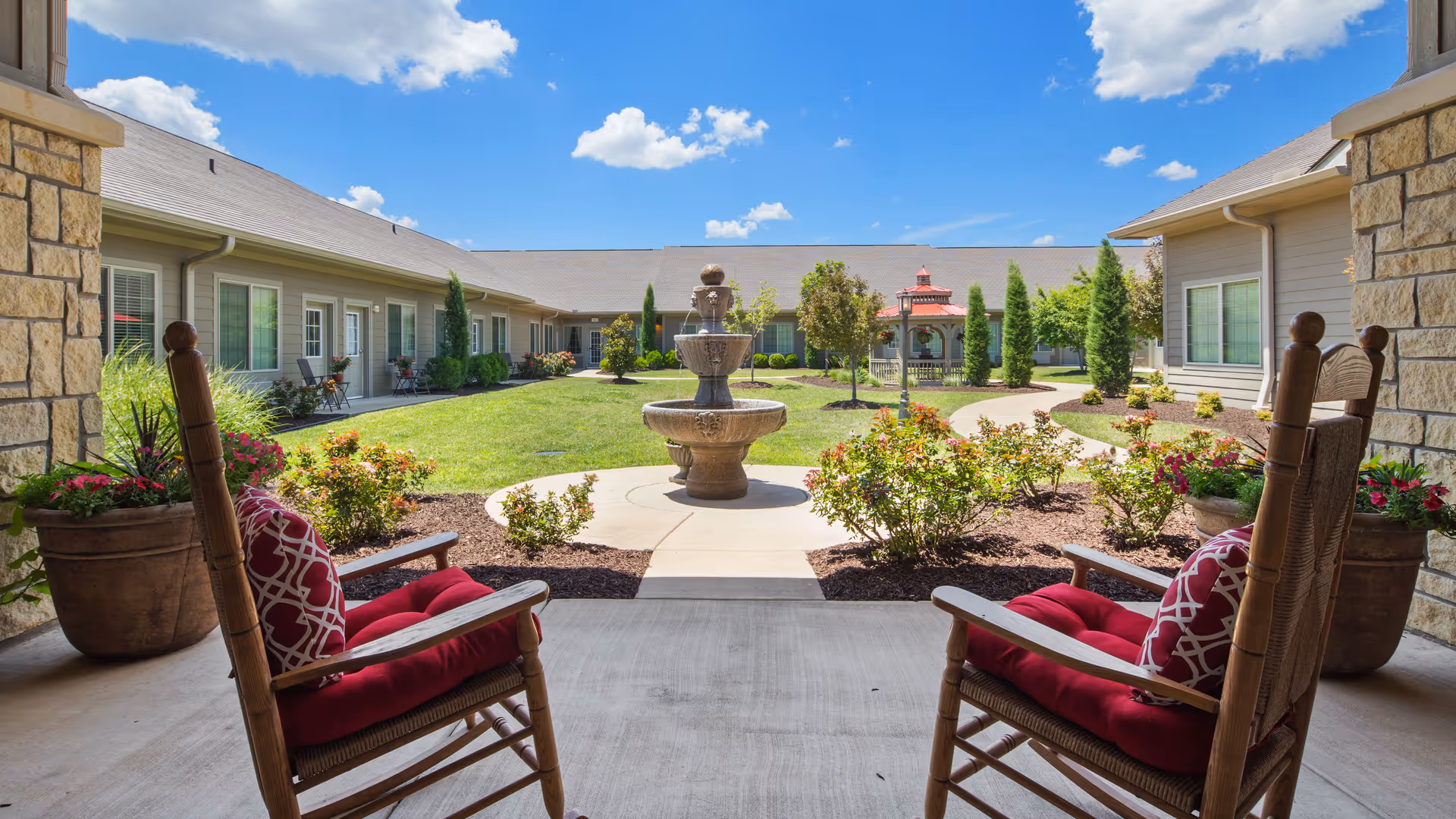 View from a covered patio area with two wooden rocking chairs featuring red cushions and patterned pillows, overlooking a landscaped courtyard with a central stone fountain, green grass, shrubs, and a gazebo in the background under a blue sky with scattered clouds.