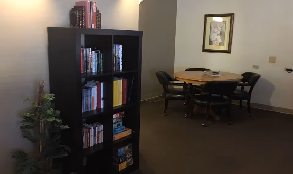 A quiet room with a black bookshelf filled with books and board games on the left side, a small green plant beside it, and a round wooden table with four black chairs on the right side. A framed picture hangs on the wall above the table.