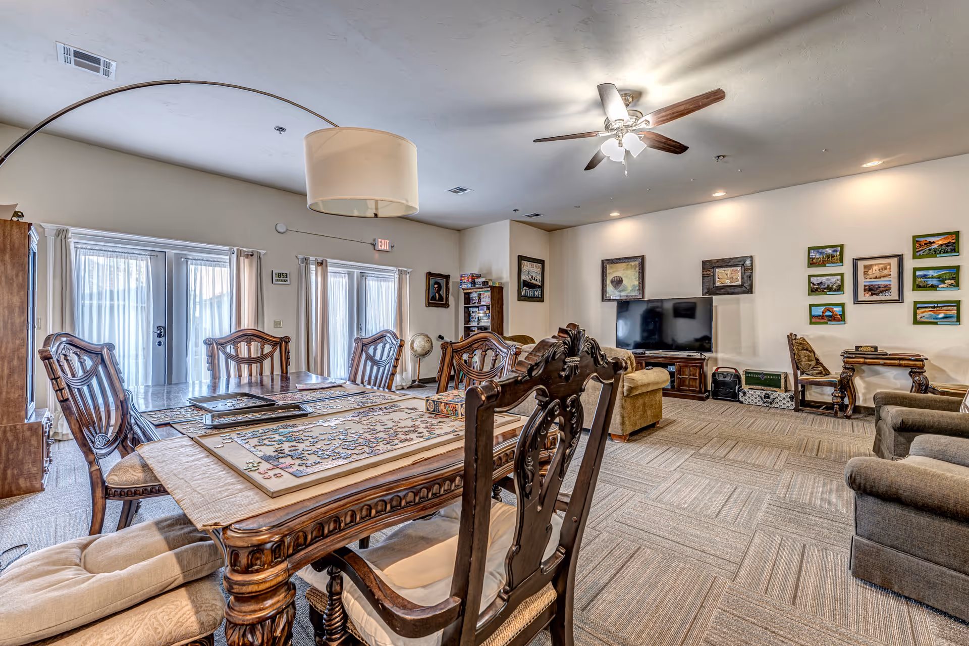 A spacious living and dining area in a senior living facility with a large wooden dining table featuring a partially completed jigsaw puzzle. The room has several ornate wooden chairs around the table, a ceiling fan with lights, a large flat-screen TV on a stand, framed pictures on the walls, and comfortable seating including armchairs and a sofa. French doors with sheer curtains allow natural light into the room.