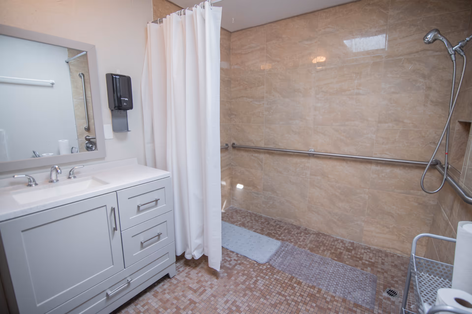A bathroom with a white vanity and sink on the left, a large mirror above the sink, and a walk-in shower area with beige tiled walls and a mosaic tiled floor. The shower has a handheld showerhead, grab bars, a white shower curtain, and a non-slip mat on the floor. There is also a metal cart with paper towels and other items on the right side.