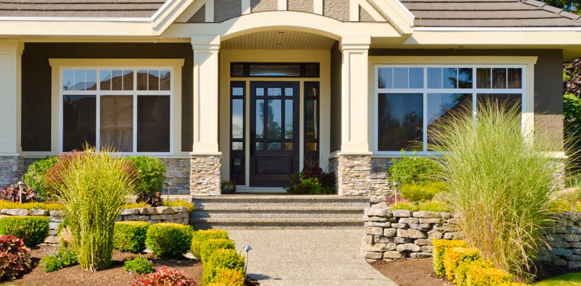 Front exterior view of a house with a covered entrance, stone pillars, large windows, and a well-maintained garden with various shrubs and plants along a stone pathway.