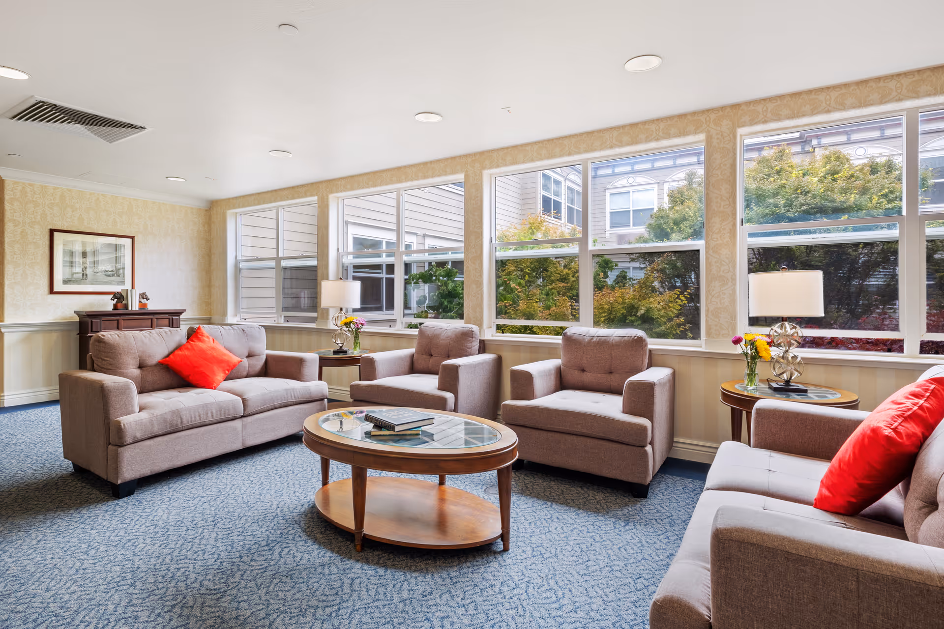 Bright communal living room with sofas and armchairs arranged around a glass-top coffee table in front of large windows.