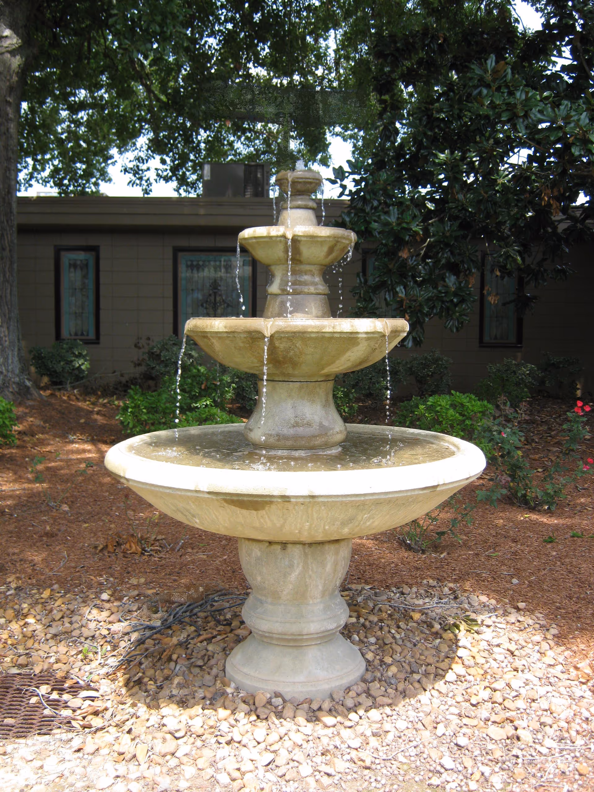 A three-tiered stone water fountain in an outdoor garden area with trees, bushes, and a building with windows in the background.