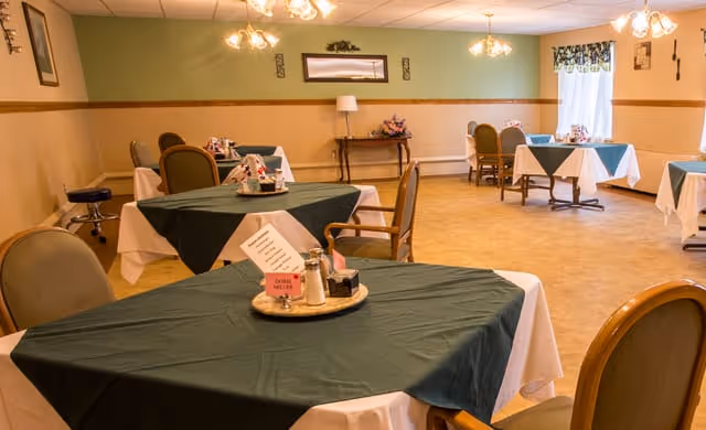 A dining room in a senior living facility with multiple tables covered with green and white tablecloths. Each table has a centerpiece with condiments and a small sign. The room has beige walls with a green accent wall, wooden chair rails, and several chairs around the tables. There are ceiling lights and a window with floral curtains letting in natural light.