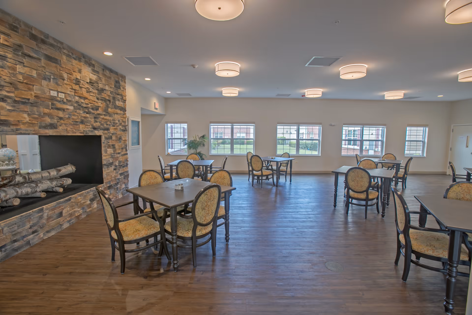 A spacious dining room with multiple tables and chairs arranged neatly. The room features a stone fireplace with logs inside on the left side, wooden flooring, and several windows letting in natural light. Ceiling lights are evenly spaced across the ceiling.