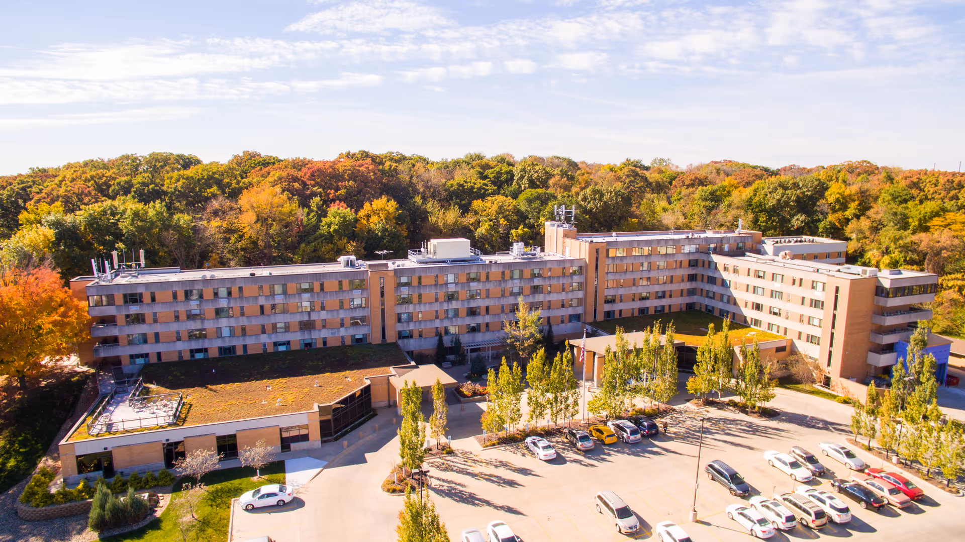 Aerial view of Meth-Wick Community, a multi-story senior living facility surrounded by trees with autumn foliage. The building has a large parking lot with several cars parked, and the sky is partly cloudy.