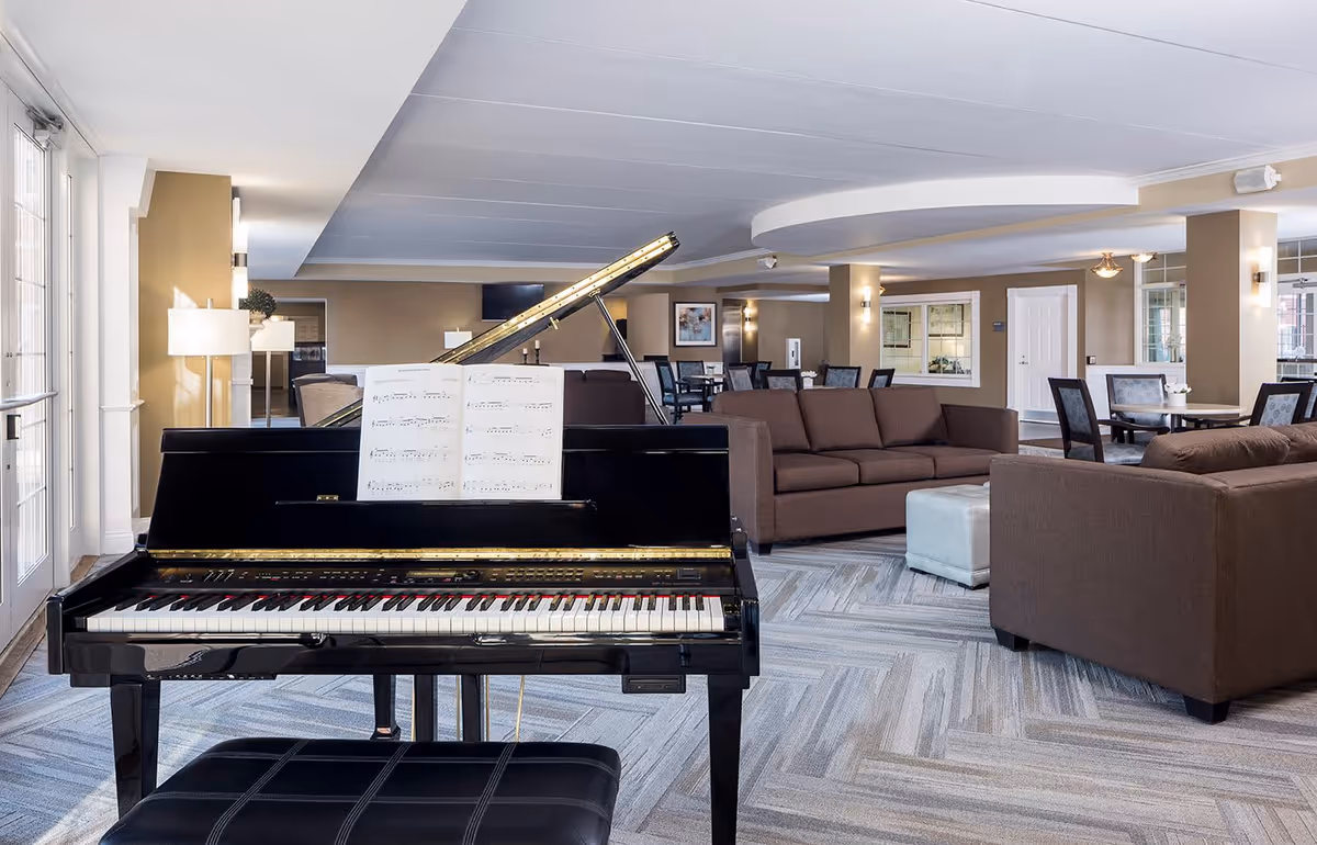 Interior view of a spacious common area in a senior living facility featuring a black grand piano with sheet music, brown sofas arranged around a white ottoman, and several dining tables with chairs in the background. The room has beige walls, large windows allowing natural light, and modern lighting fixtures.