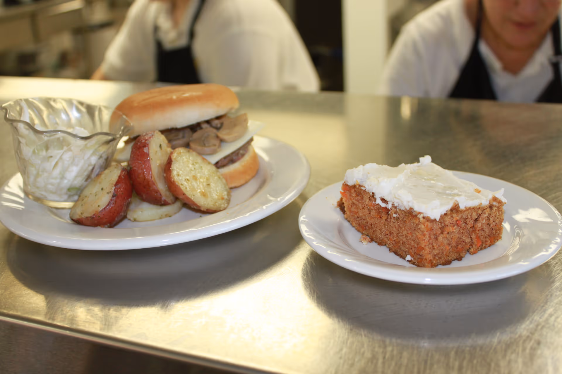 A meal served on a metal counter with a sandwich containing mushrooms and cheese, roasted potato slices, a small bowl of coleslaw, and a slice of frosted carrot cake on separate white plates. Two people in white shirts and black aprons are partially visible in the background.