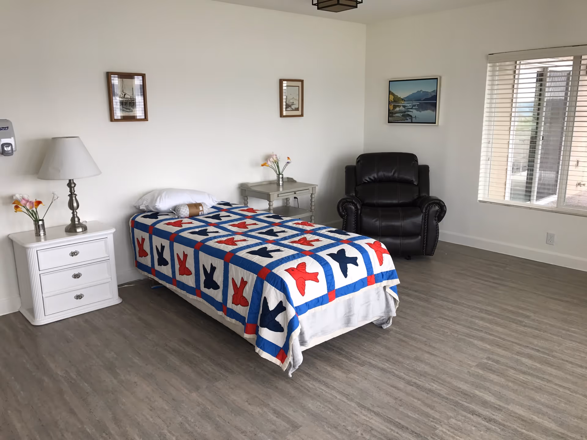 A simple bedroom with a single bed covered by a colorful quilt featuring red, blue, and white patterns. The room has light-colored walls and flooring. There is a white nightstand with a lamp and a small vase of flowers on the left side of the bed, and a gray side table with flowers on the right side. A black leather armchair is positioned near a window with white blinds. Three framed pictures hang on the walls.