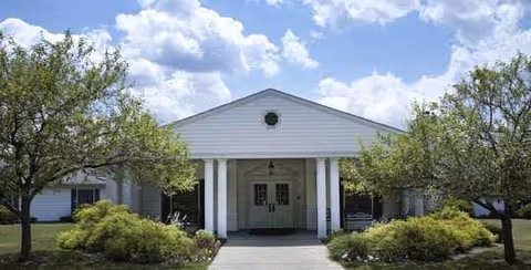 Front view of a white, columned senior living facility entrance with landscaping.