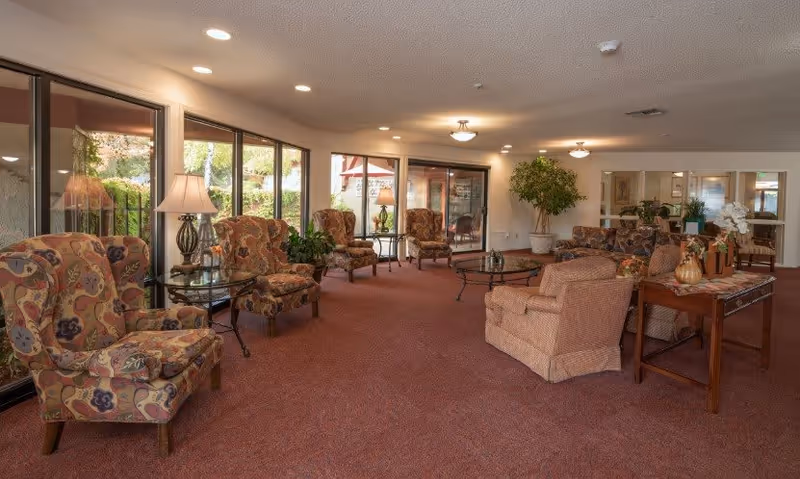 A spacious senior living common area with patterned armchairs, a sofa, glass-top tables, lamps, and large windows overlooking an outdoor garden. The room is carpeted in a reddish tone and decorated with plants and flowers.