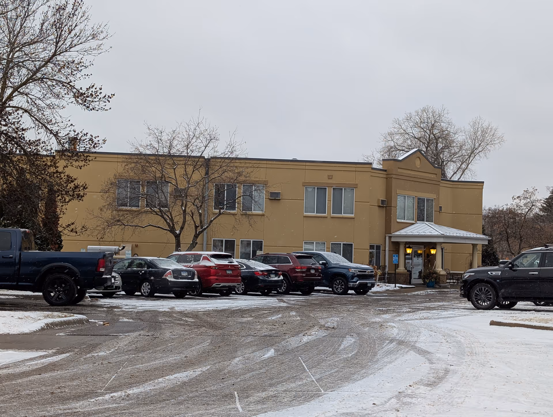 Exterior view of a two-story beige building with several windows and a covered entrance. There are multiple cars parked in front of the building on a snowy day, with bare trees around the parking area.
