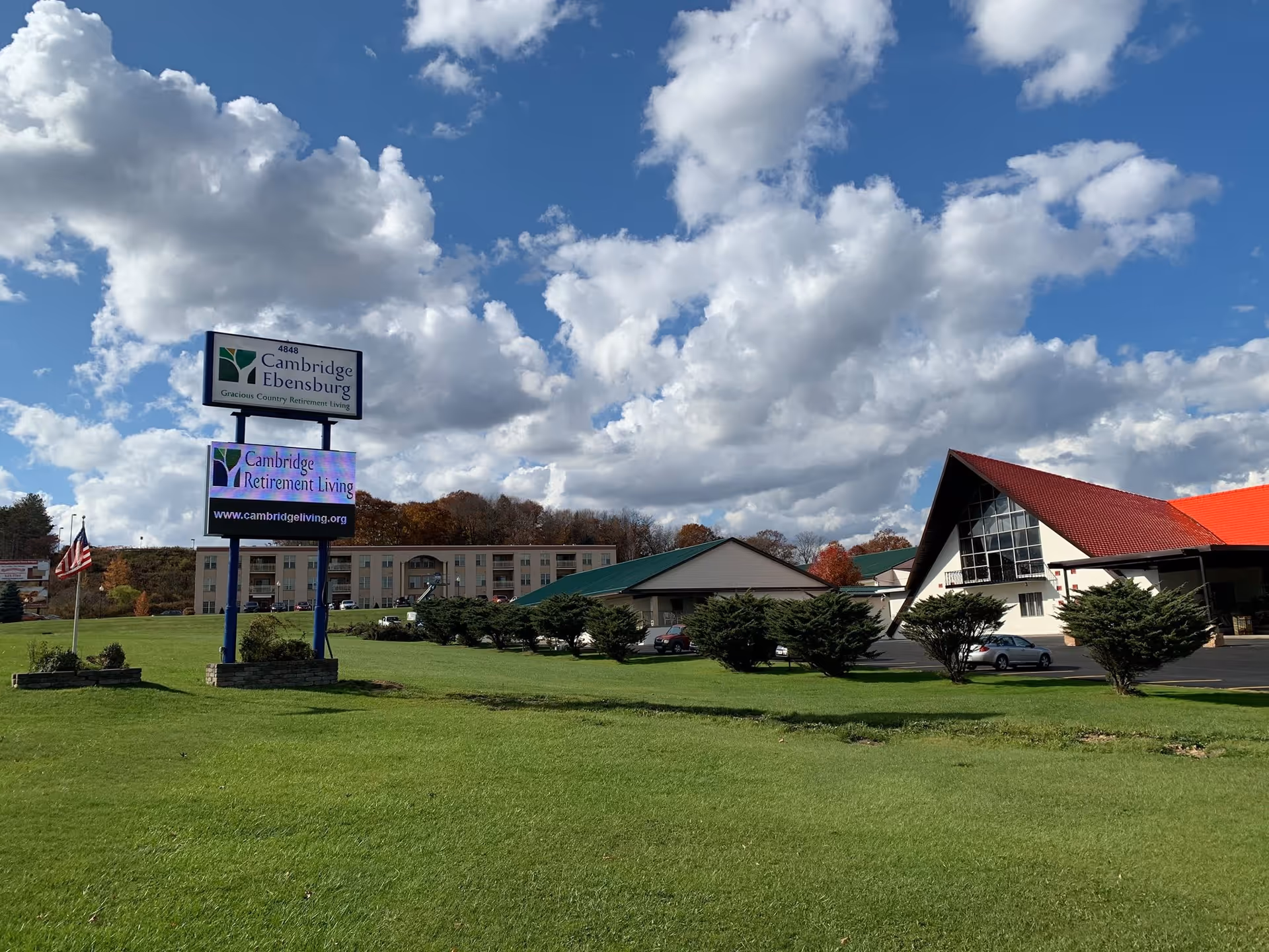 Exterior view of Cambridge Ebensburg Senior Living facility with a large green lawn in the foreground, a sign displaying the facility name and website, several buildings including one with a red roof, and a partly cloudy blue sky.