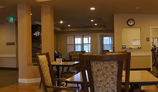 Interior view of a common area in an assisted living facility with wooden chairs and tables, beige walls, a clock on the wall, and windows letting in natural light.
