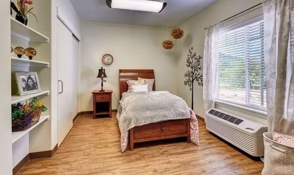 Bright single-occupancy bedroom with a wooden bed, nightstand and lamp, shelving, a wall tree decal, large window and air conditioner unit.