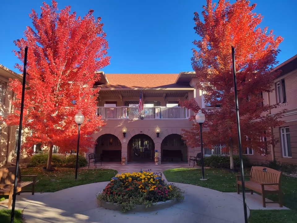 Courtyard of a residential building with bright red autumn trees, a central flowerbed, benches, and an arched entrance under a clear blue sky.