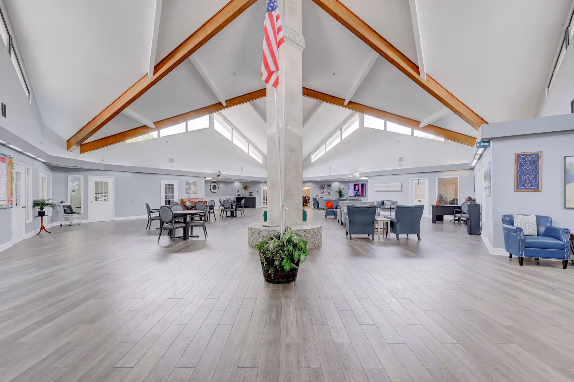 Spacious interior common area with high vaulted ceilings featuring wooden beams and large windows near the ceiling. The room is divided by a central pillar with an American flag hanging from it and a potted plant at its base. On the left side, there are round tables with chairs, and on the right side, there are several armchairs and sofas arranged around small tables. The walls are light blue with various framed artwork and a television mounted on the far right wall.