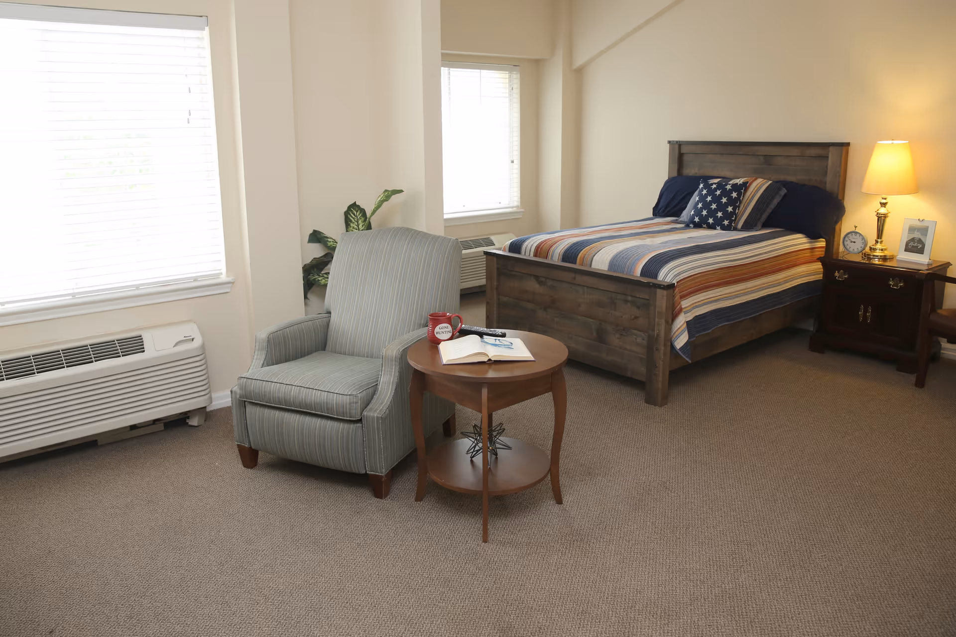 A cozy bedroom with a wooden bed featuring a striped bedspread and star-patterned pillow, a wooden nightstand with a lamp, clock, and picture frame, a comfortable armchair next to a small round wooden table with an open book and a red mug, two windows with blinds, and a potted plant in the corner.