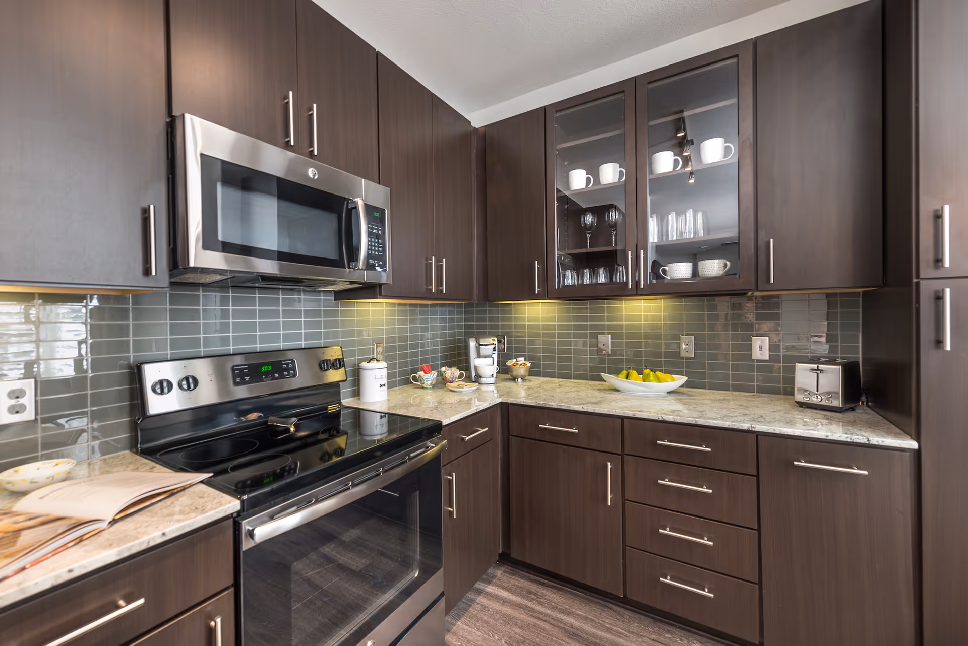 Modern kitchen with dark wood cabinets, a stainless steel microwave and stove, a gray tiled backsplash, and a granite countertop holding a coffee maker, a bowl of green pears, a toaster, and some cups and glasses in a glass-front cabinet.