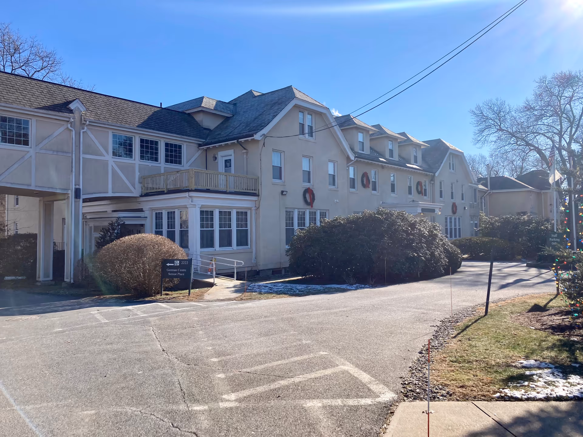 Exterior view of a large beige building with multiple windows and a sloped roof under a clear blue sky. The building has holiday wreaths on the walls and a small balcony. There are bushes and a paved driveway in front, with a sign indicating 'German Centre Senior Place' and another sign for staff parking. Some snow patches are visible on the ground.