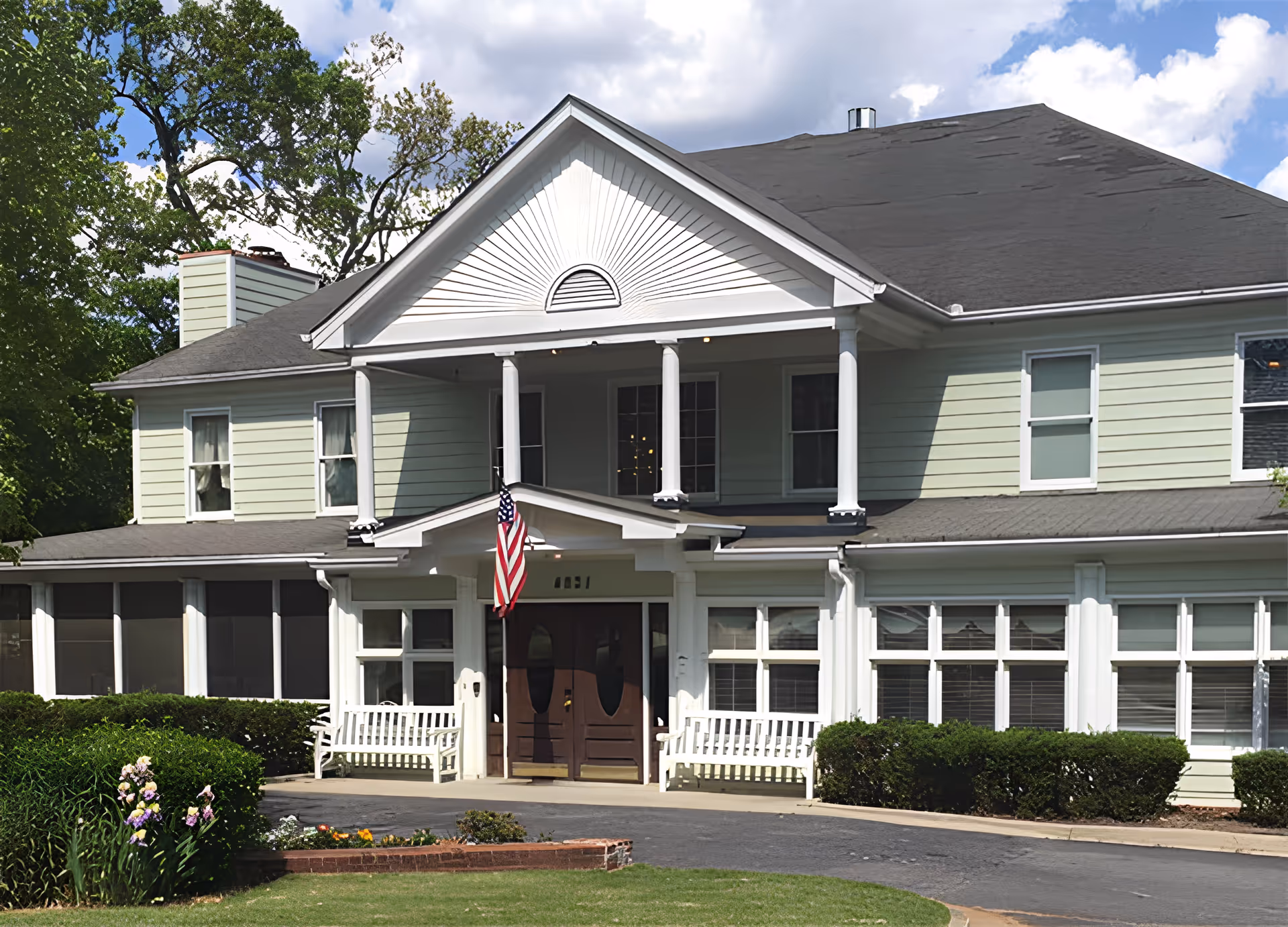 Front exterior of a two-story pale green senior living building with a columned entrance, American flag, benches, and landscaped front lawn.