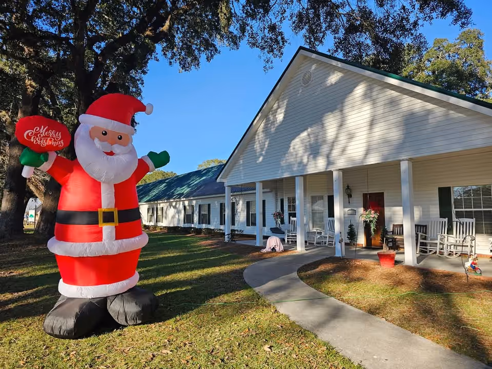 Large inflatable Santa on the lawn in front of a single-story senior living building with a covered porch and rocking chairs.