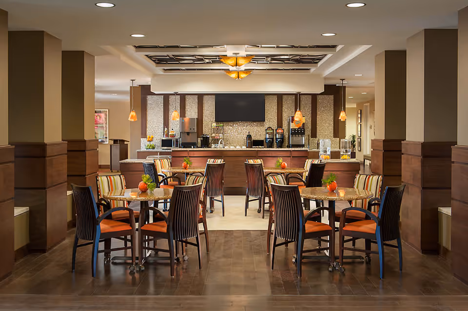 A well-lit dining area with several round tables and chairs featuring orange cushions and striped backs. Each table has a small decorative plant. In the background, there is a counter with beverage dispensers, a microwave, and hanging pendant lights. The room has a warm color palette with wood paneling and a decorative ceiling light fixture.