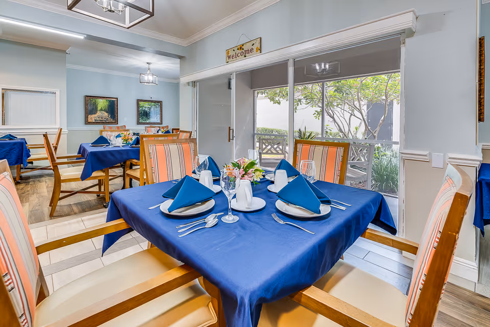 Bright dining room with tables set in blue linens and napkins, chairs, and a large window overlooking a garden.