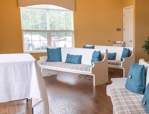 Interior room with white wooden benches featuring plaid cushions and blue pillows, a table covered with a white cloth, large window with blinds showing parked cars outside, and hardwood flooring.