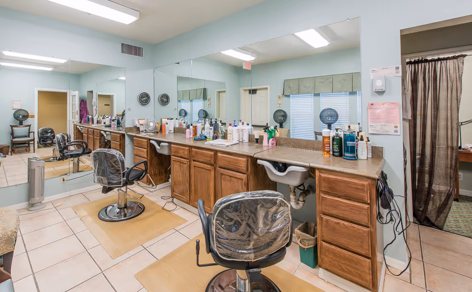Salon area with styling chairs in front of a mirrored wall, a countertop full of hair products and a small sink.