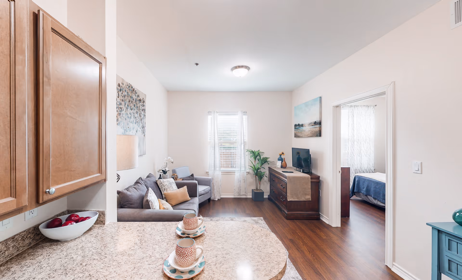 A bright and cozy living room area with a gray sofa, an armchair, and decorative pillows. A wooden cabinet with a TV and a plant is against the far wall. The room has a window with sheer curtains letting in natural light. To the right, there is a doorway leading to a bedroom with a bed covered in a blue blanket. The foreground shows a granite countertop with two cups and saucers, and wooden kitchen cabinets are visible on the left.
