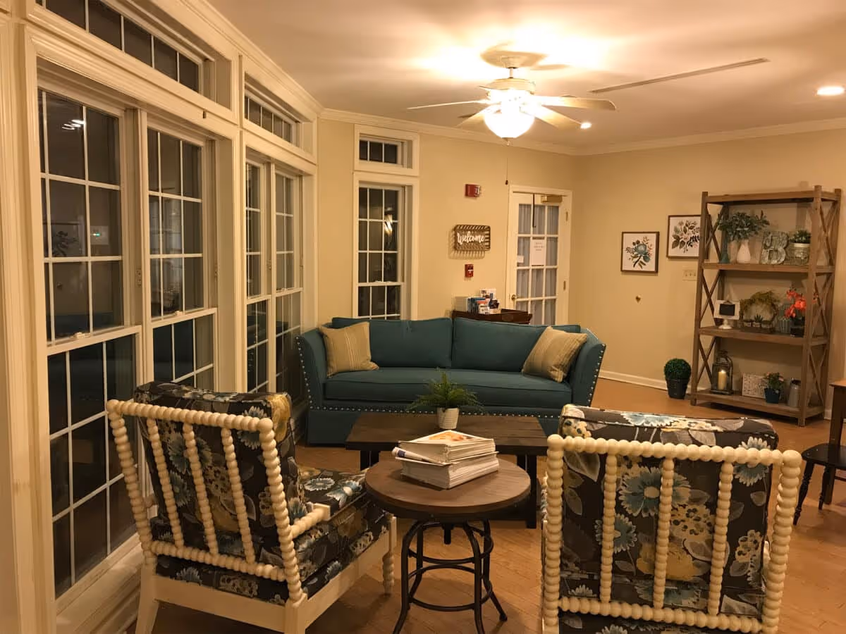 Cozy living room with a teal sofa, two patterned armchairs around coffee tables and a shelving unit beside large multi-pane windows.