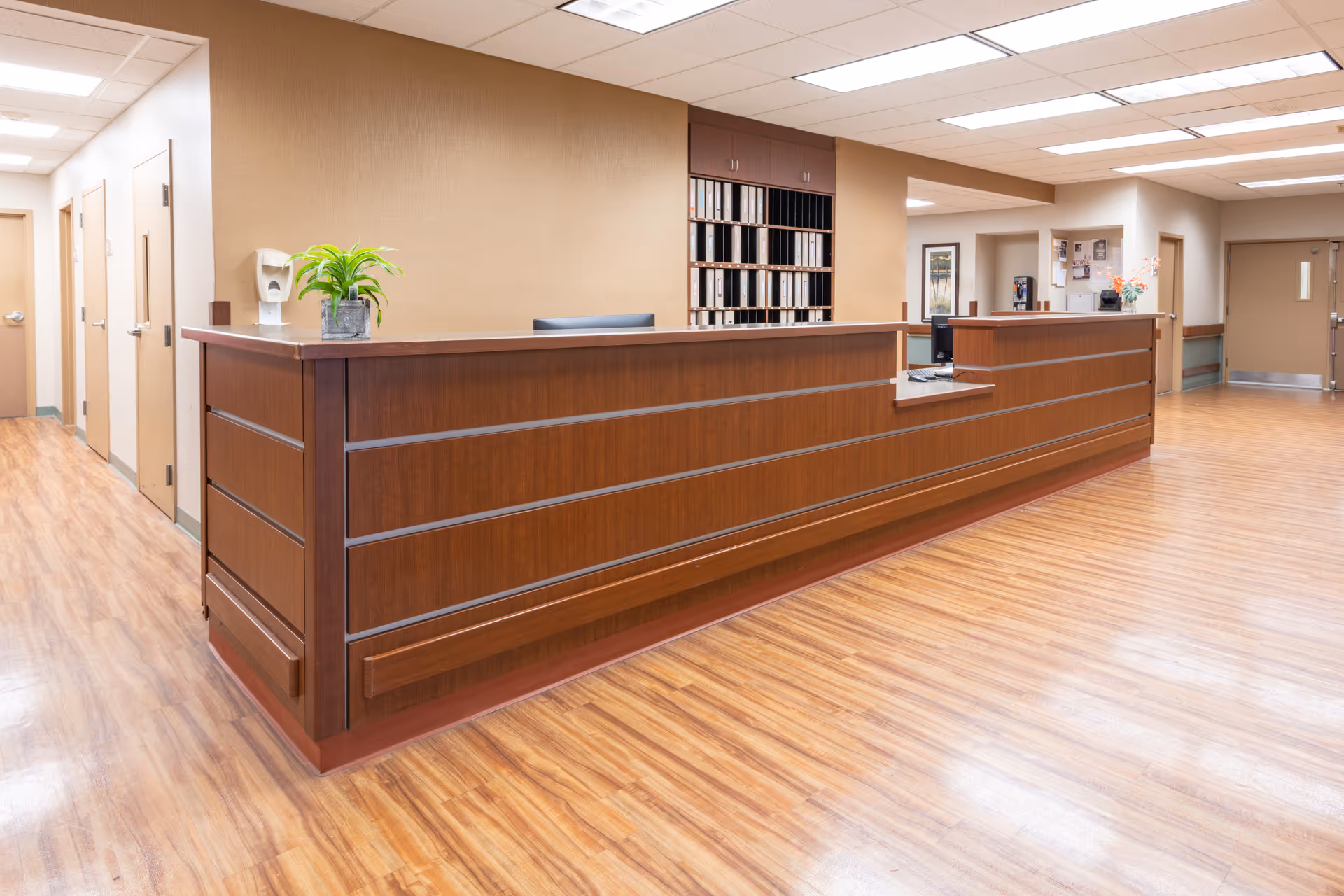 A spacious reception area with a long wooden front desk, wood-patterned flooring, beige walls, and fluorescent ceiling lights. Behind the desk are shelves with organized files. There is a small green plant on the desk and a hand sanitizer dispenser mounted on the wall to the left. Several doors and a hallway are visible in the background.