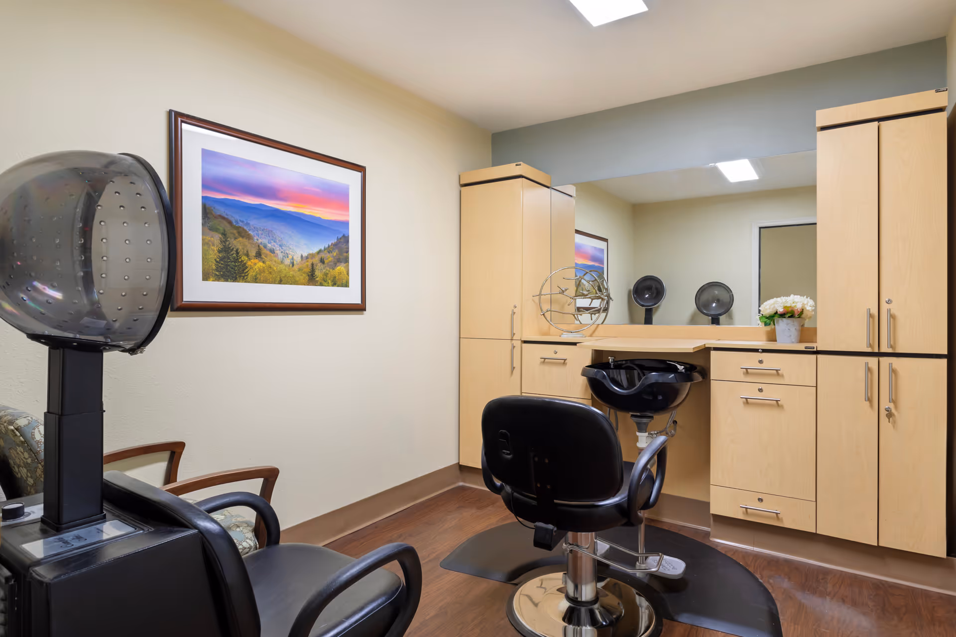 Interior of a hair salon area in a senior living facility with a black salon chair, a hair dryer, a black hair washing sink, light wood cabinets, a large mirror, and a framed landscape picture on the wall.
