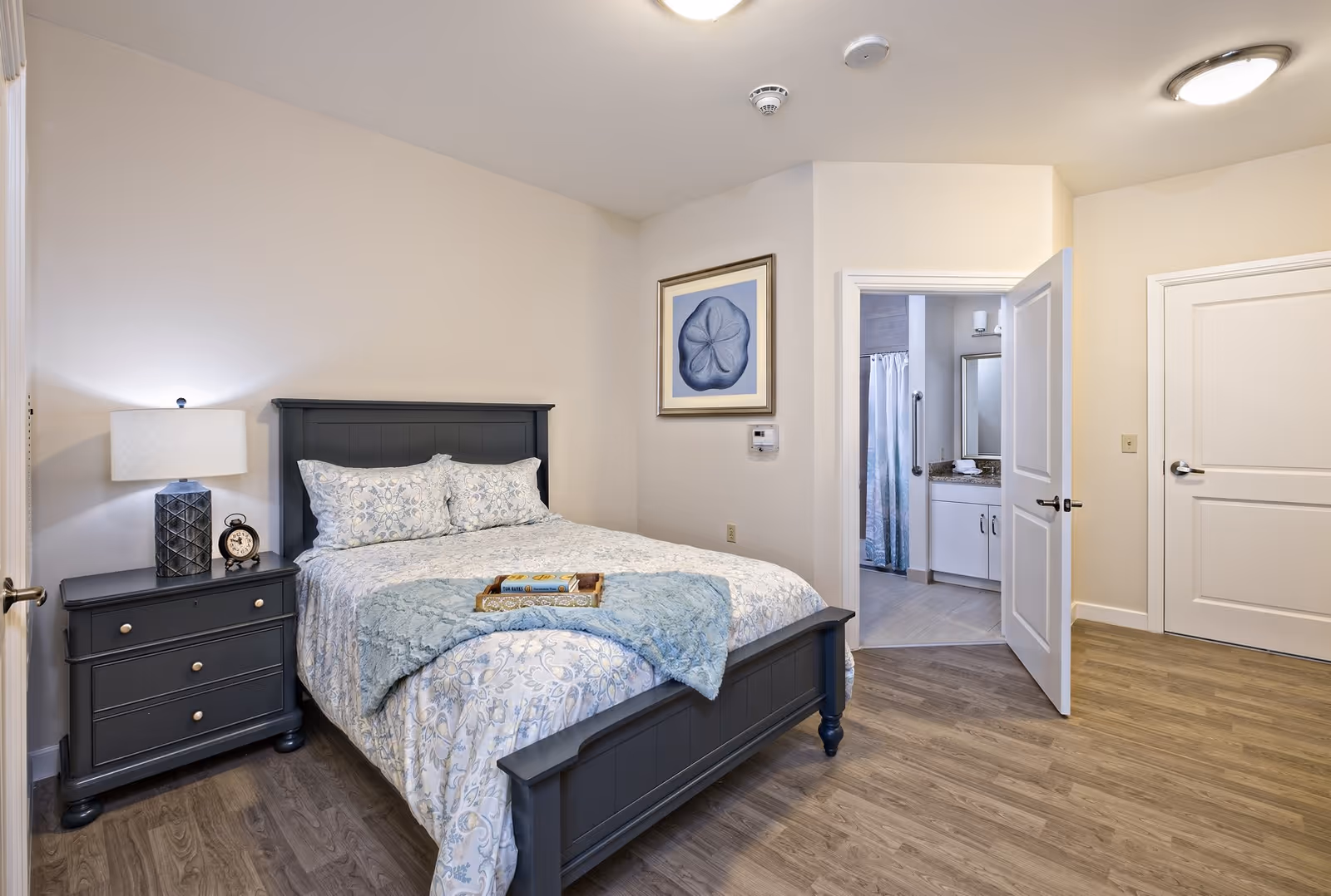 A cozy bedroom with a dark wooden bed frame and matching nightstand. The bed is made with light-colored floral bedding and a blue throw blanket. On the nightstand, there is a lamp and a small clock. The room has light-colored walls and wood flooring. A framed blue artwork hangs on the wall above a thermostat. An open door reveals a bathroom with a granite countertop, white cabinets, and a shower curtain.
