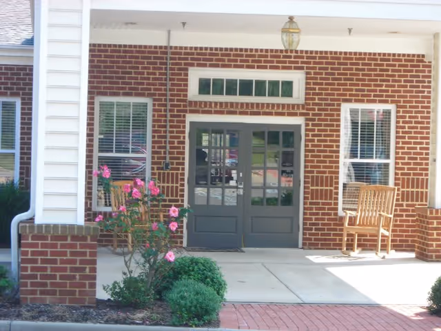 Entrance to a brick building with double gray doors and windows on either side. There are two wooden chairs on the porch, a hanging light fixture above the doors, and some pink flowers and green shrubs in front of the entrance.