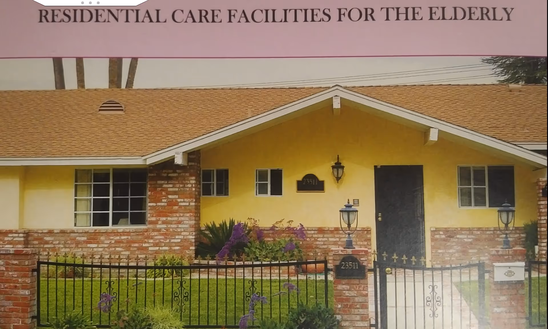 Front exterior view of a single-story residential care facility for the elderly with a yellow facade, brick accents, a brown shingle roof, black front door, two black lantern-style lights, a black metal fence, and a well-maintained garden with purple flowers.