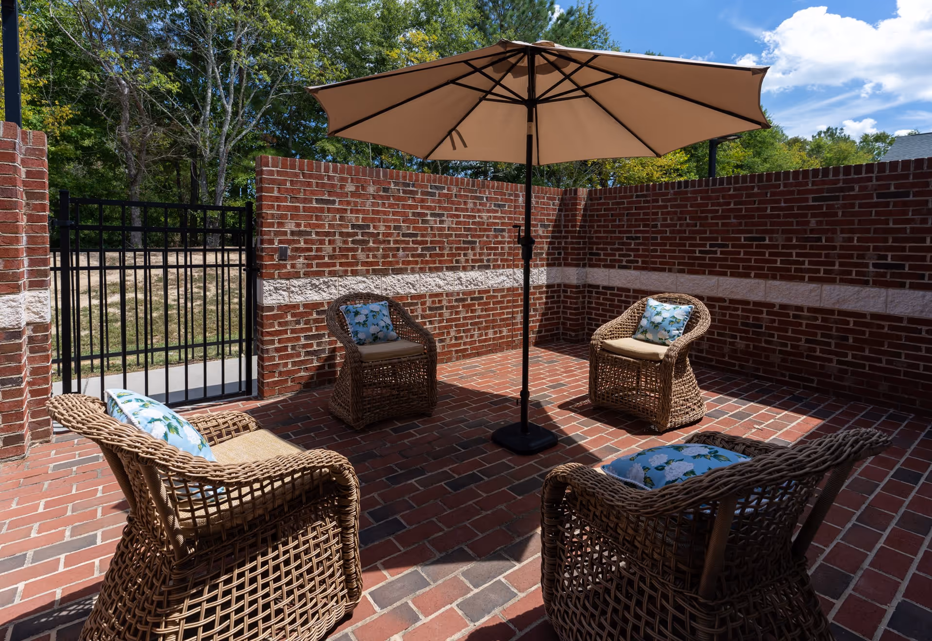 Outdoor patio area with four wicker chairs arranged around a large beige umbrella. The patio is enclosed by red brick walls with a black metal gate leading outside. The chairs have cushions with a floral pattern. Trees and blue sky are visible beyond the brick enclosure.